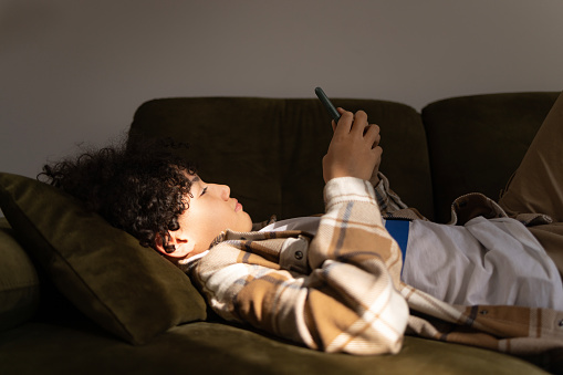 African American boy text messaging on smart phone while relaxing on sofa in the living room.
