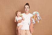 Indoor shot of stressed tired mother holding her little baby daughter, doing cosmetic procedures in the morning, looking at camera, posing isolated over brown background.