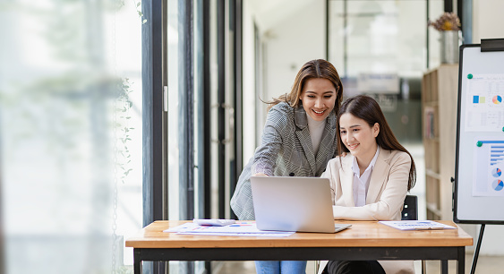 Two beautiful Asian businesswoman in conversation Exchange ideas at work Company employees working together by talking and guiding each other with papers, graphs and desk laptops.