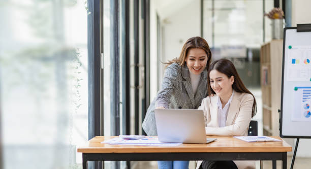 Two beautiful Asian businesswoman in conversation Exchange ideas at work Company employees working together by talking and guiding each other with papers, graphs and desk laptops. stock photo