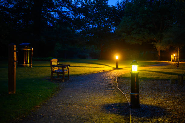 A park at night. A stony path cuts across a lawn with a park bench and is illuminated by electric lights. The lamps cast a yellow light and long shadows.