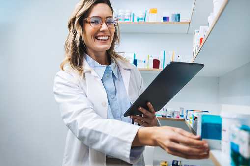 Happy pharmacist getting medication from a shelf in a chemist. Female healthcare worker filling prescriptions using a tablet. Woman working in a pharmacy.