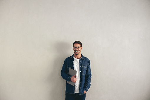Happy young businessman smiling while standing against a grey wall with a laptop. Cheerful young businessman wearing business casual and eyeglasses in a modern office.