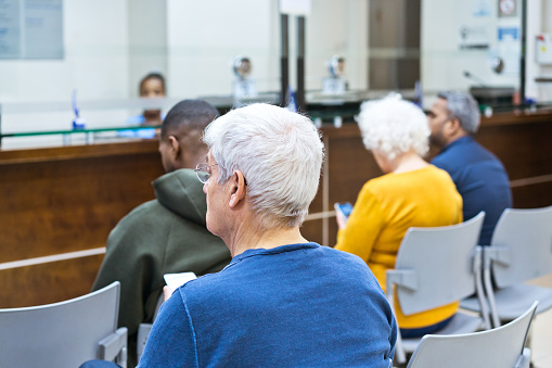Back view of multiracial group of people sitting in waiting room in hospital. Reception in the background.
