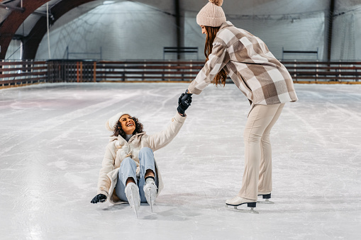 Two beautiful young female friends ice skating in the ice skating ring indoors. Young woman helping her friend get up after falling in ice skate rink.