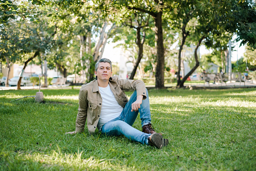 Portrait of gray haired man in a park with positive expression sitting on the grass looking at the camera.