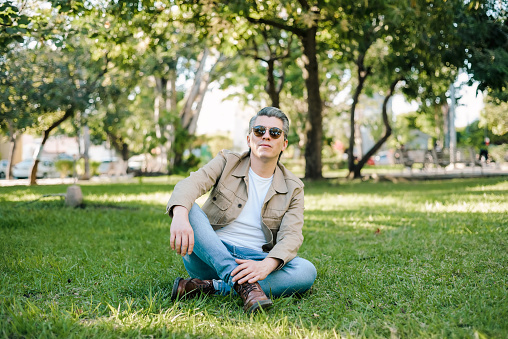 Portrait of gray-haired man wearing sunglasses in a park sitting on the grass looking at the camera.