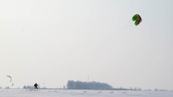 man in black pulling a green kite in the field,man snowkiting in the field. Kitesurfer ride the parachute in the wind among the snow.