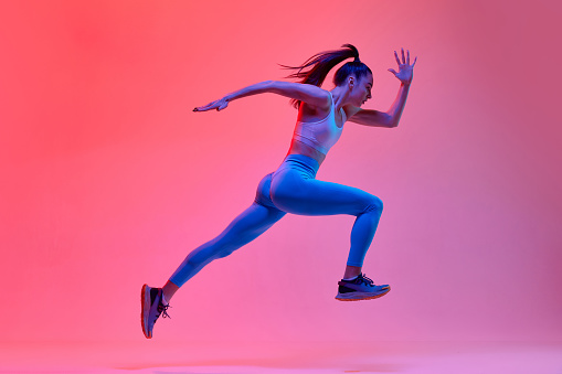 Dynamic portrait of young active girl, athlete, runner in motion, training over pink studio background in neon light. Concept of sportive lifestyle, health, endurance, action and motion. Ad