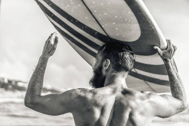istock Young hipster man with surfboard on his head greeting friends with a gesture - Guy having fun doing extreme sport - Sporty bearded man training with surfboard on the sea - Black and white editing 1470757059