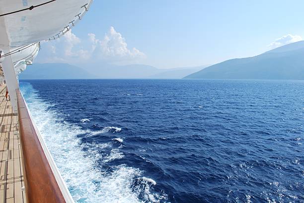 huge white colored cruise ship next to a large sailboat in front of an island with mountain range, view from distance, water with lots of small waves in front