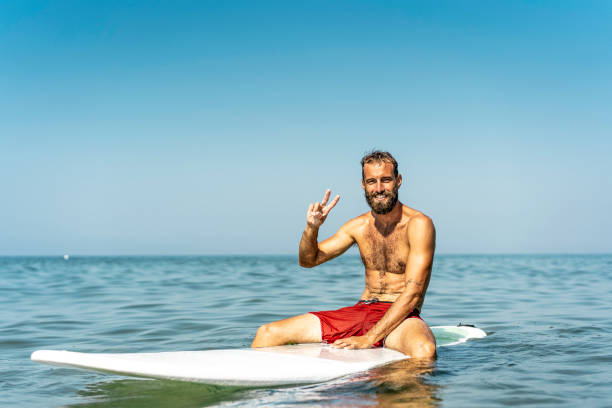 młody hipster siedzący na desce surfingowej witający przyjaciół gestem zwycięstwa - facet dobrze się bawiący uprawiając sporty ekstremalne - sportowy brodaty mężczyzna trenujący z deską surfingową na morzu - koncepcja wolności - co surfing zdjęcia i obrazy z banku zdjęć