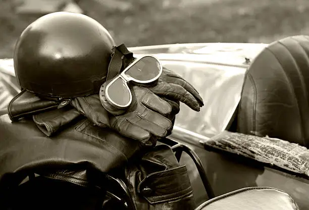 Vintage motorcycle leathers, helmet and goggles placed on a vintage motorcycle seat with sidecar in the background.