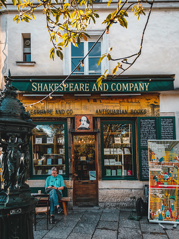 The photo shows the exterior of Shakespeare and Company Bookstore on the early morning and a man reading a book while sitting, located on a busy street in the heart of Paris. The building is a charming, old-fashioned structure with rustic wooden beams and an ornate black-and-white sign above the entrance that reads 