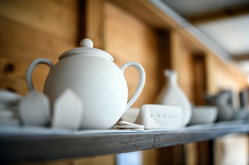Three shelves with porcelain and ceramics dishes, tea pots and mugs standing by the wall.