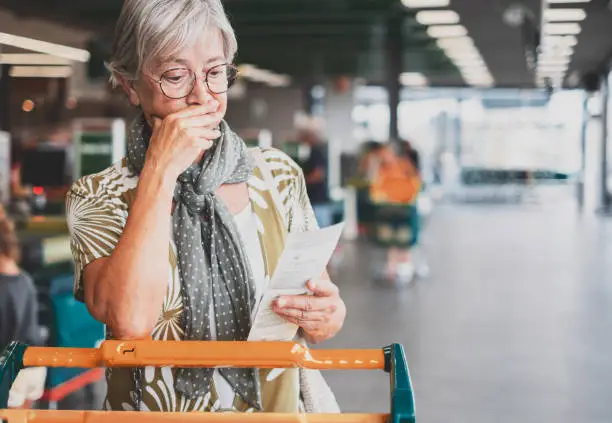 Photo of Senior woman in the supermarket checks her grocery receipt looking worried about rising costs - elderly lady pushing shopping cart, consumerism concept, rising prices, inflation