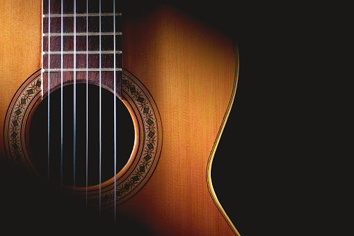 A serious senior man sits at home with his granddaughter.  She holds a guitar and looks down as he reaches forward to point to a fret.