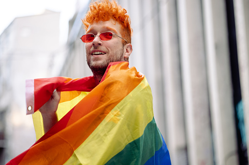 Young man holding a rainbow flag on the street. Alternative looking man covered with a rainbow flag
