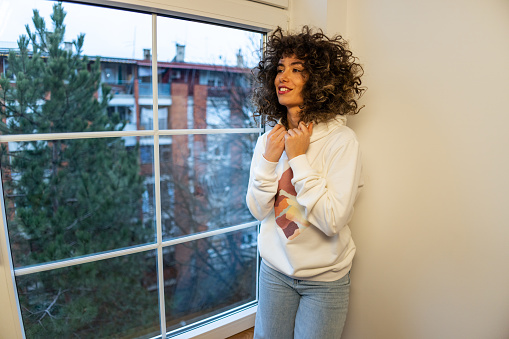 Portrait of a Caucasian young beautiful girl with curly hair wearing a white hood with application while standing near window