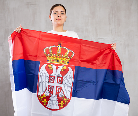Contented young woman with Serbia flag in hands posing proudly against light unicoloured background