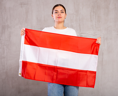 Confident patriotic young woman in casual wear holding flag of Austria against gray wall indoors