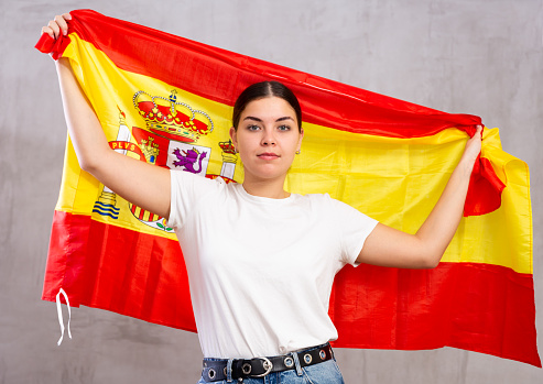 Serious young woman in casual wear holding Spanish flag against gray wall background indoors