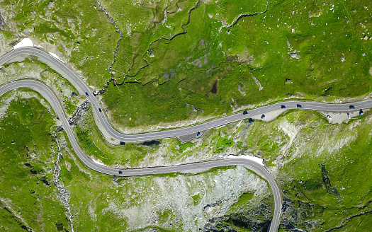 Aerial view above a mountain road winding on the alpine pastures. The asphalt road is transited by cars passing the mountain. A stream flows along the road. Carpathia, Romania.