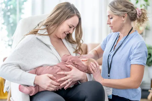 A new mother sits with her infant on her lap as she breast feeds her.  A female lactation consultant is overseeing the feeding to ensure proper posture for the mother and latch of the baby.