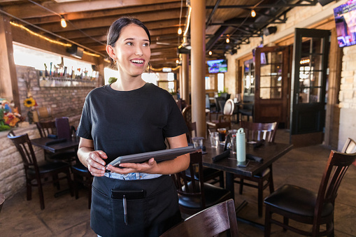 The young adult waitress smiles at her unseen coworker who is asking her a question as she stands with the digital tablet on the patio of the restaurant.