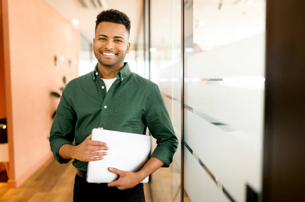 working with joy. smiling cheerful indian businessman wearing casual green shirt standing with laptop computer in office - minority professional occupation business ethnic imagens e fotografias de stock