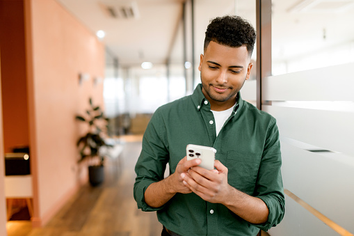 Guapo hispano está usando un teléfono inteligente para enviar mensajes, enviar mensajes de texto, joven de cabello oscuro con elegante camisa verde casual usando teléfono inteligente photo