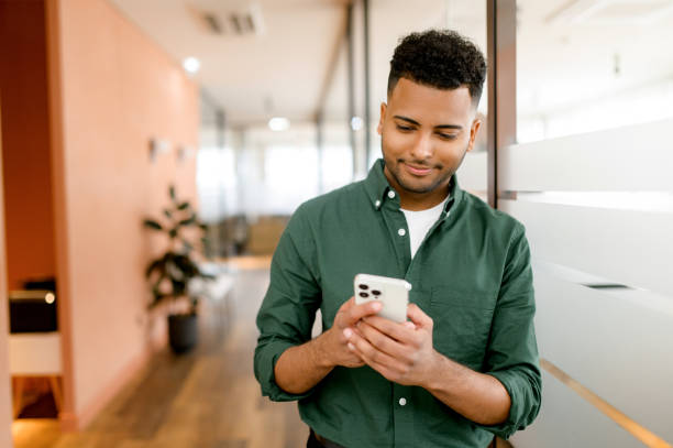 Handsome hispanic guy is using smartphone for messaging, texting, young dark-haired man in smart casual green shirt using smartphone stock photo