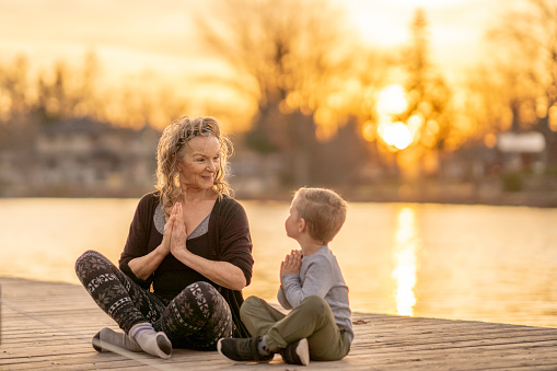 A senior woman sits cross legged outside with her Grandson beside her as they focus on their breathing and meditate.  They are both dressed comfortably and are focused on their relaxation as they sit lakeside