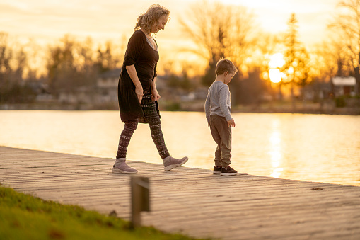 A senior woman walks along the water with her Grandson as they spend quality time together at sunset.  They are both dressed casually and are looking out over the water.