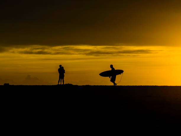 istock Sunrise silhouettes - lovers and a lone surfer heading out 1470809628