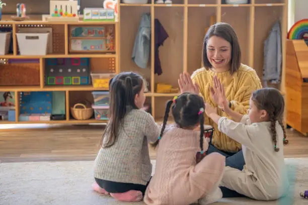 A small group of Kindergarten students sit on the floor with their teacher as they each hold a set of bells sing along together.  They are each dressed casually and are focused on shaking their instruments to the beat.