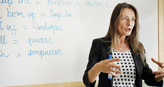 An ethnic female graduate teaching assistant stands at a large whiteboard and writes down Spanish verbs to conjugate for her students. The shot is over the shoulder of several students.