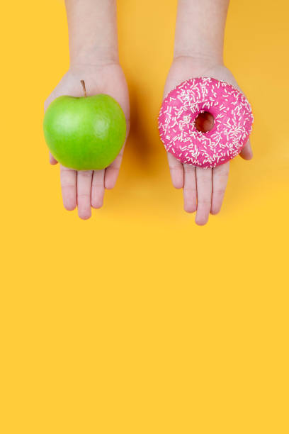 des mains féminines tenant une pomme verte et un beignet rose. concept d’aliments sains et d’habitudes de vie par rapport à une alimentation et à un mode de vie malsains. fond jaune. espace de copie. - healthy eating diabetes dieting healthcare and medicine photos et images de collection