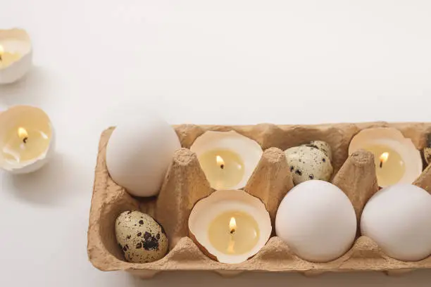Easter holiday concept. Candlights in eggshell. White chicken and quail eggs in paper tray on white background. Creative approach. Copy space