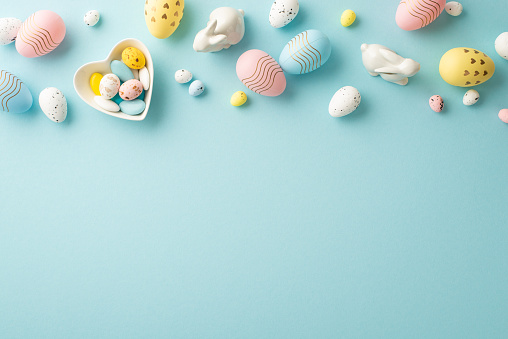 Easter decorations concept. Top view photo of colorful easter eggs heart shaped saucer with sweets and ceramic bunnies on isolated pastel blue background with copyspace