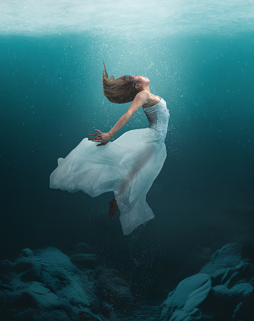 Beautiful girl in artistic dance underwater with flowing dress and natural lighting from ocean surface.