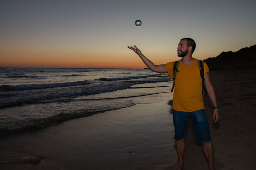 Male traveler in casual clothes with backpack tossing crystal sphere while standing on wet beach near waving sea in evening