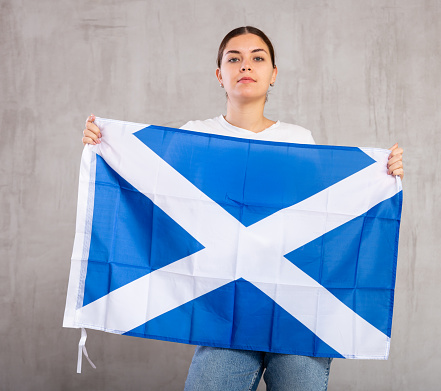 Serious patriotic young woman waving state flag of Scotland against gray wall background