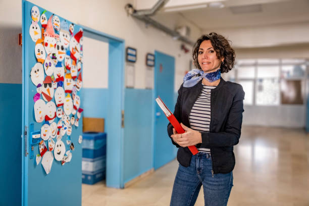 Caucasian female smiling elementary teacher with brown short curly hair, standing at school corridor and looking at the camera. Portrait view. She is holding a red file and wearing a dark jacket. Horizontal indoors shot with copy space. Back to school concept.