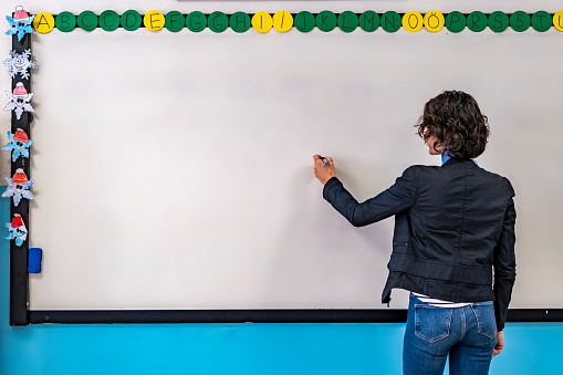 Waist up shot of fit woman with brown short curly hair standing in front of white board and writing. Casual rear view. Horizontal indoors shot, copy space. Back to school concept.