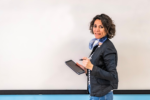 Waist up shot of Caucasian female smiling teacher with brown short curly hair, looking at camera and holding a note pad and pencil. Casual portrait view. Horizontal indoors shot, copy space. Back to school concept.