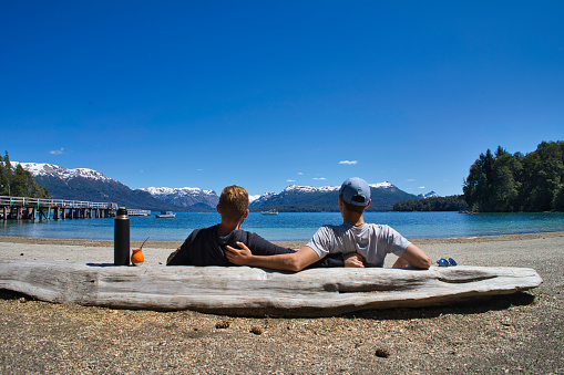 Two men hugging and leaning on a fallen log enjoy drinking mate and gazing at the lake pier and mountains on a sunny day.