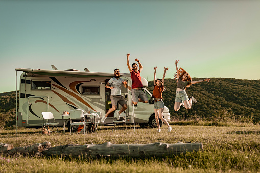 Group of cheerful friends having fun while jumping with raised arms during camping day by the trailer. Copy space.