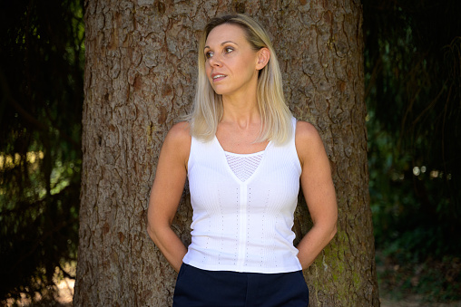 Cute thoughtful trendy blond middle aged woman posing against a tree looking aside with a smile