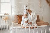 Happy mothers day concept. A little girl repeats after her beautiful mother self-care after a shower, sitting in white bathrobes and with a towel on her head in a cozy bedroom at home